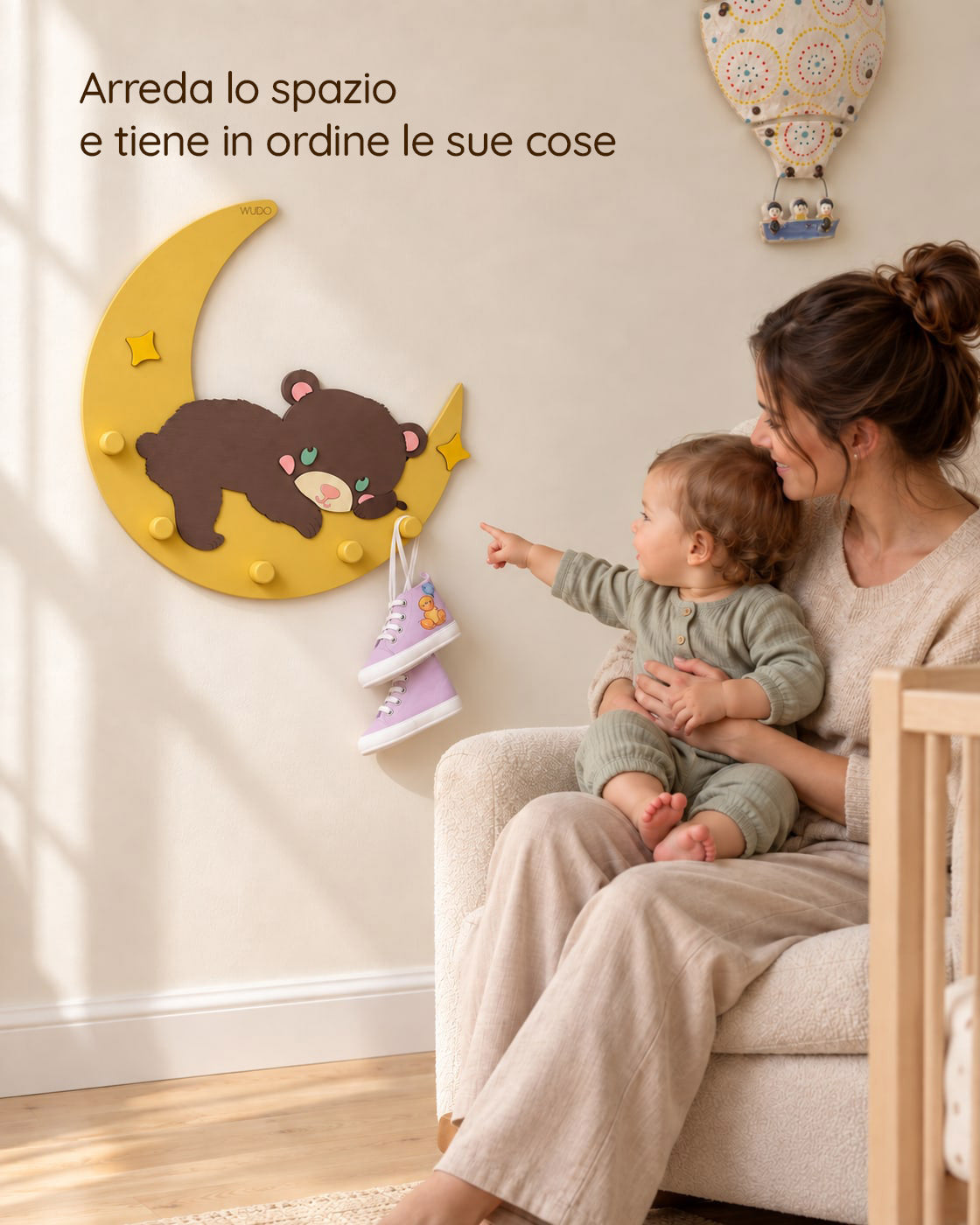 Mamma seduta in cameretta con il bambino in braccio che indica l'appendiabiti Mielo con l'orsetto sulla luna, con la scritta: Arreda lo spazio e tiene in ordine le sue cose