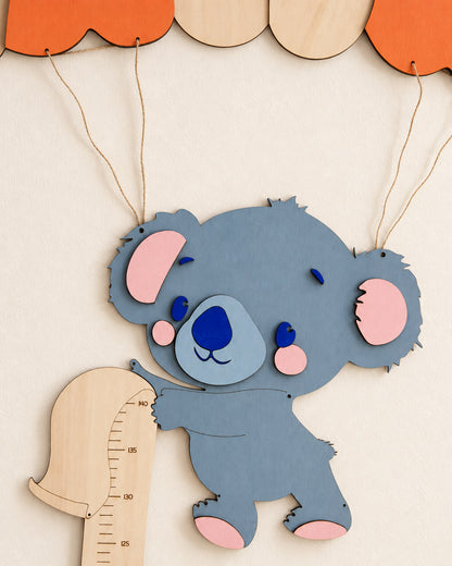 Koala in legno dipinto azzurro e rosa appeso con lo spago al paracadute, accanto alla scala da 125 a 140 cm